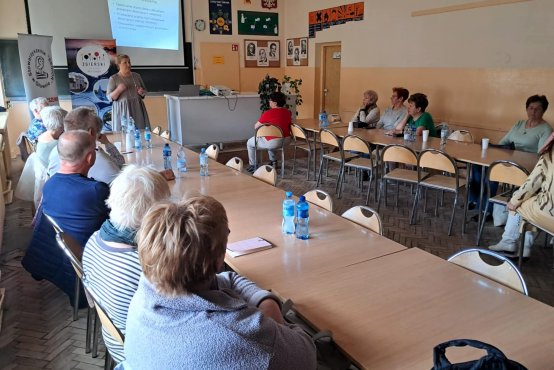 W jasnej sali przy dwóch długich stołach siedzą kobiety i mężczyżni, pośrodku stoi prawniczka prowadząca zajęcia, w tle na ścianie prezentacja. 